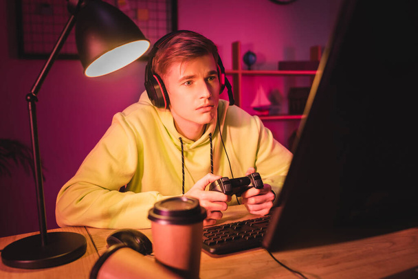 KYIV, UKRAINE - AUGUST 21, 2020: Focused player in headset using joystick near takeaway coffee and computer on blurred foreground  - 写真・画像