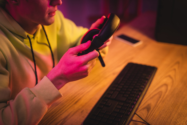 Cropped view of gamer holding headset near computer keyboard on blurred background  - Foto, afbeelding