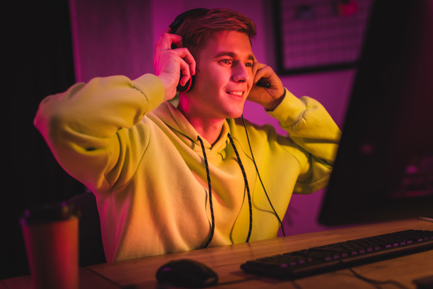 Smiling gamer wearing headset near computer and coffee to go on blurred foreground  - Photo, image