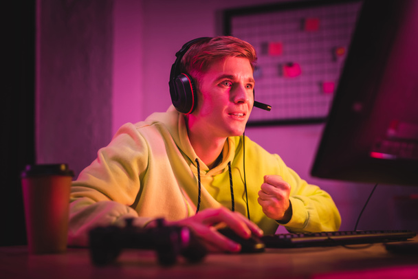 Gamer a headset mutat igen gesztus használata közben számítógép közelében joystick és kávé menni elmosódott előtérben - Fotó, kép