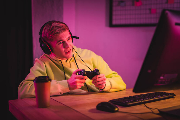 KYIV, UKRAINE - AUGUST 21, 2020: Focused gamer using joystick while playing video game on computer near coffer to go on blurred foreground  - Фото, зображення