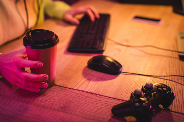 KYIV, UKRAINE - AUGUST 21, 2020: Cropped view of joystick near gamer holding takeaway coffee and using computer keyboard on blurred background  - Photo, image