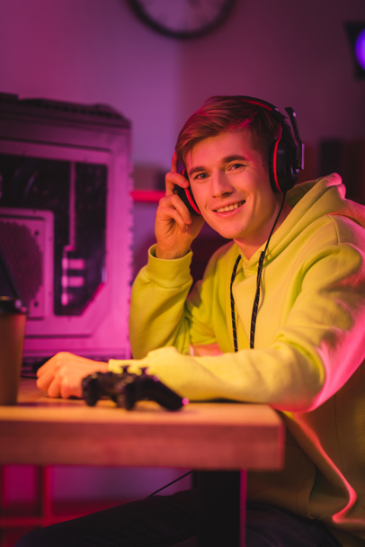 Smiling gamer holding headphones near joystick and coffee to go on blurred foreground  - Fotó, kép