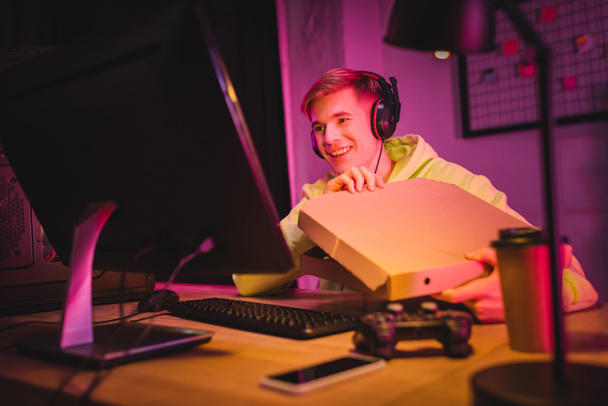 Smiling gamer in headphones holding pizza box near computer, joystick and smartphone on blurred foreground  - Photo, Image