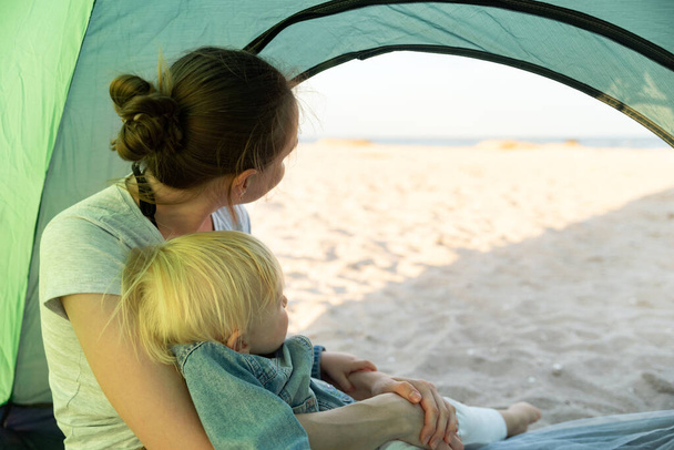 小さな赤ちゃんと母は、観光テントの中に座っている。キャンプ場からの海とビーチの景色. - 写真・画像