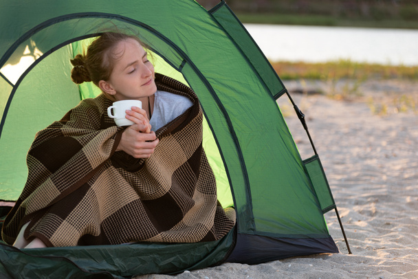 Mujer joven en la tienda bebe té de la mañana. Vacaciones de camping en campo. - Foto, Imagen
