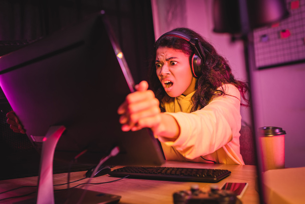 Őrült afro-amerikai játékos headset tartja számítógép monitor kávé közelében, okostelefon és joystick elmosódott előtérben  - Fotó, kép