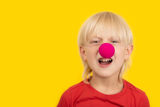 Boy with clown nose discontentedly looking at camera and making faces. Portrait close up on yellow background. - Photo, Image