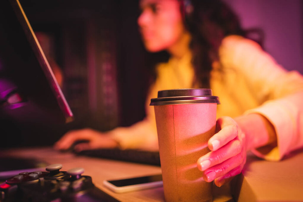 KYIV, UKRAINE - AUGUST 21, 2020: Coffee to go near joystick and gamer using computer on blurred background  - Foto, afbeelding
