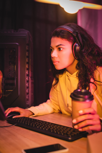 Afro-Amerikaanse speler in headset spelen van video game op de computer en het houden van take-away koffie op wazig voorgrond  - Foto, afbeelding