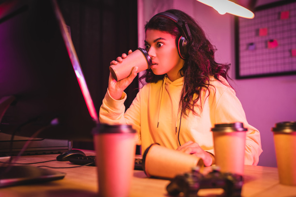 Innoissaan afrikkalainen amerikkalainen gamer kuulokkeet juominen kahvia ho tietokoneen lähellä ja joystick hämärtynyt etualalla  - Valokuva, kuva