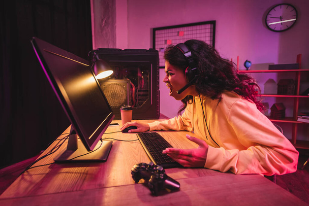 KYIV, UKRAINE - AUGUST 21, 2020: Angry african american gamer in headset using computer near gamepad and coffee to go  - 写真・画像