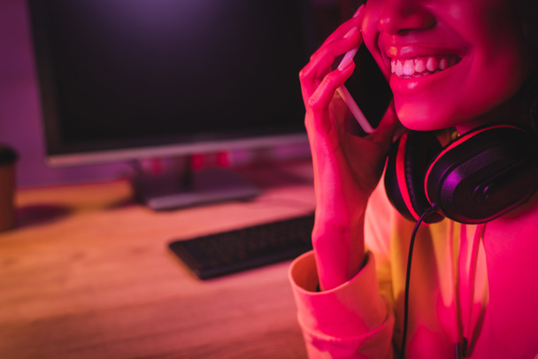 Vágott kilátás mosolygós afro-amerikai nő fülhallgató beszél okostelefon közelében számítógép elmosódott háttér  - Fotó, kép