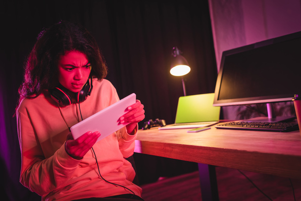 Concentrado afroamericano gamer en auriculares usando tableta digital cerca de dispositivos en fondo borroso  - Foto, Imagen