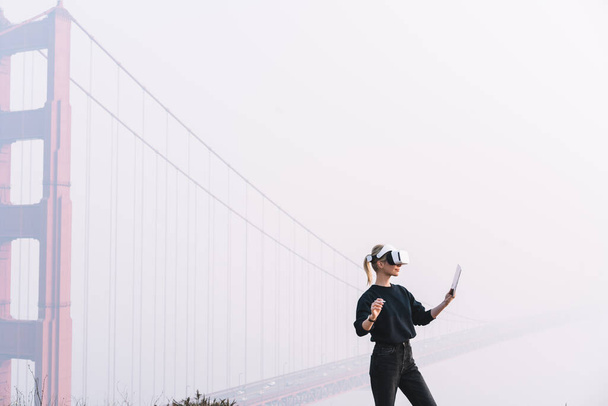 Side view of focused female in casual clothes standing near Golden Gate in VR device during free time in city - Фото, зображення