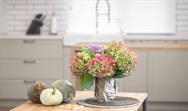 Composición otoñal de flores de colores de hortensias y calabazas sobre un fondo borroso del interior de una cocina moderna y luminosa de cerca. - Foto, imagen