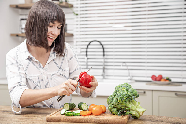 Cheerful brunette girl cuts vegetables on salad on the background of modern kitchen interior. - Photo, Image
