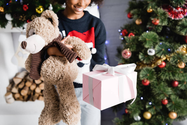 cropped view of smiling african american girl holding gift box and teddy bear on blurred background - Foto, Imagen