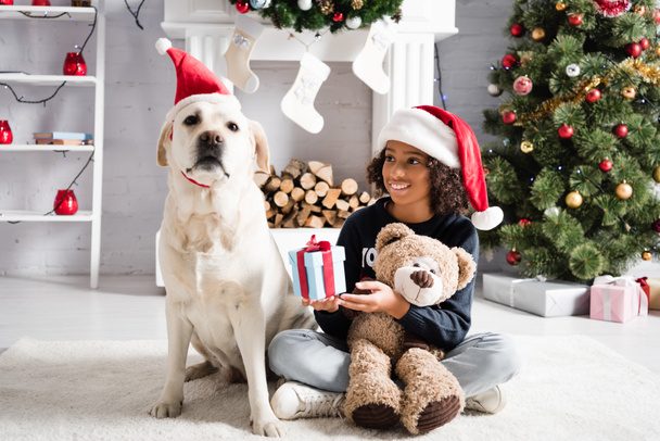 happy african american girl holding gift box and teddy bear while sitting on floor near labrador dog on blurred background - Foto, Imagem