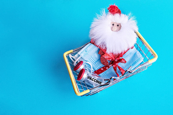 In un cestino della spesa in miniatura si trovano maschere protettive mediche legate con un nastro festivo e Babbo Natale con il vaccino coronavirus su sfondo blu. Capodanno e il concetto di Natale in pandemia. - Foto, immagini