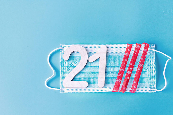 Номер 21 и праздничная лента на медицинской защитной маске на синем фоне. Концепция Рождества и Нового года в условиях пандемии коронавируса. - Фото, изображение