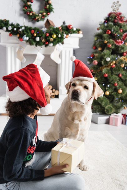african american girl in santa hat holding gift box while sitting near labrador dog on blurred background - Photo, image