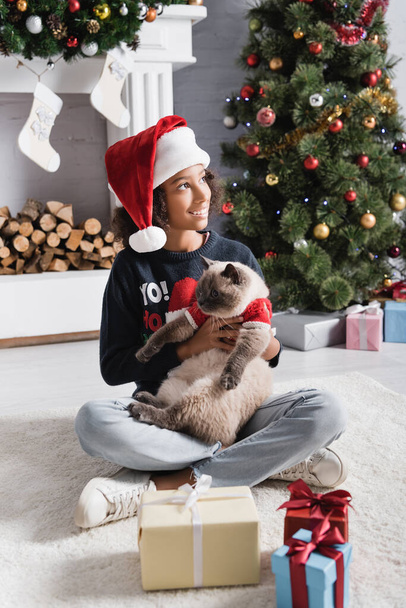 happy african american girl in santa hat looking away while sitting near christmas tree and holding fluffy cat - Photo, Image