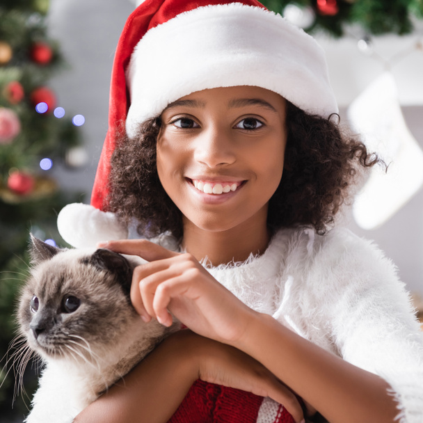 fröhliche afrikanisch-amerikanische Mädchen mit Weihnachtsmütze streichelt Katze und schaut in die Kamera auf verschwommenem Hintergrund - Foto, Bild