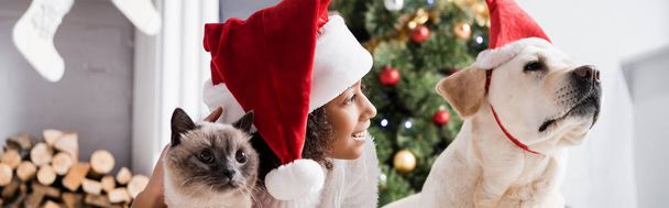 fille afro-américaine souriante en chapeau de Père Noël près du chien labrador et chat moelleux sur fond flou, bannière - Photo, image