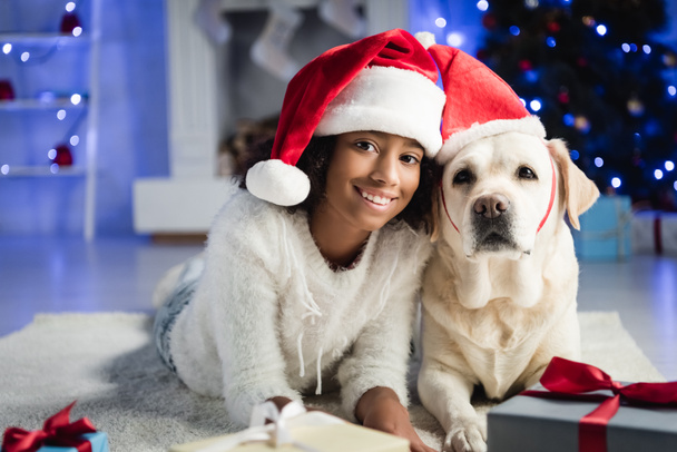joyeuse fille afro-américaine et chien labrador couché sur le sol dans chapeaux Santa sur fond flou - Photo, image