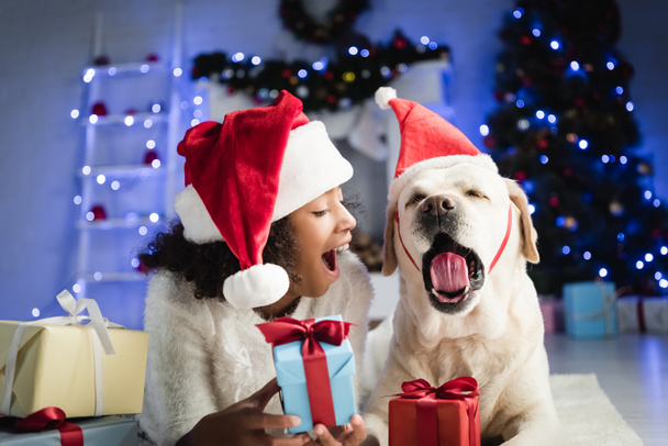 african american girl and labrador in santa hat yawning while lying on floor near gift boxes on blurred background - Fotó, kép