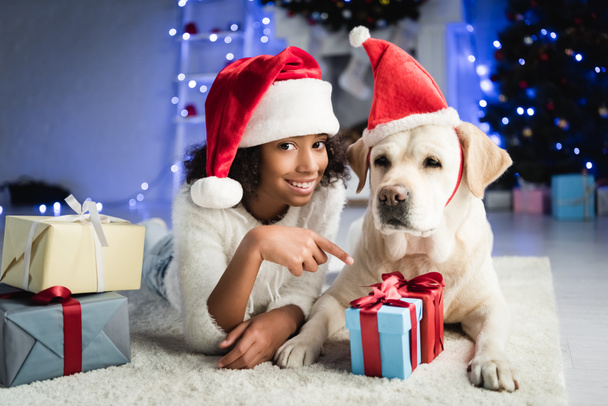 fröhliches afrikanisch-amerikanisches Mädchen zeigt mit dem Finger auf Geschenkbox, während sie auf dem Boden neben Labrador-Hund auf verschwommenem Hintergrund liegt - Foto, Bild