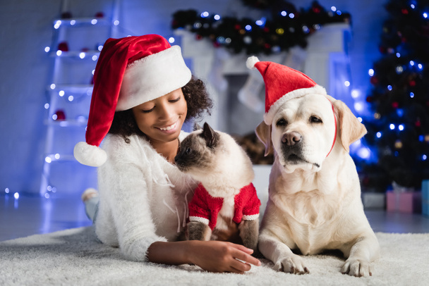 joyful african american girl lying on floor near labrador and cat on blurred background with christmas decoration - Photo, Image
