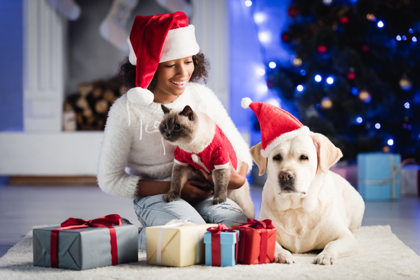 felice ragazza afroamericana in cappello di Babbo Natale, abbraccio gatto e seduto vicino labrador sul tappeto bianco con regali, su sfondo sfocato - Foto, immagini