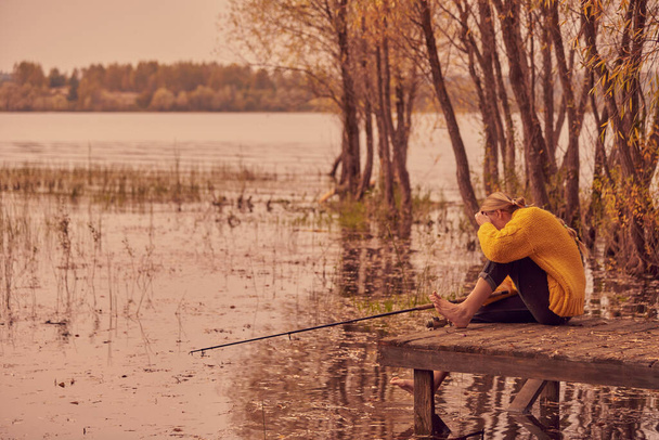 A barefoot Girl in a warm sweater sits with a fishing rod on a wooden bridge by the lake. Autumn evening landscape. - Fotó, kép