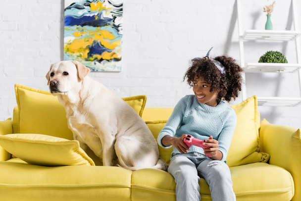 KYIV, UKRAINE - OCTOBER 02, 2020: Smiling african american girl playing with joystick and looking at retriever, while sitting on sofa at home - Фото, зображення