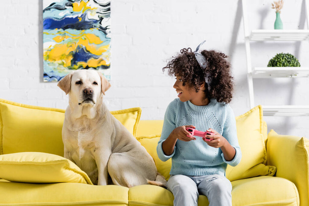 KYIV, UKRAINE - OCTOBER 02, 2020: excited african american girl with joystick looking at labrador, while sitting on sofa at home - Фото, зображення