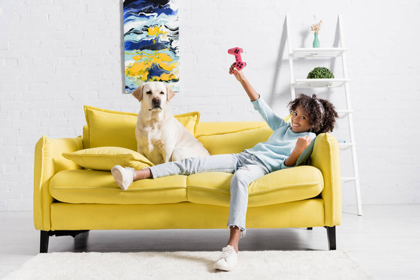 KYIV, UKRAINE - OCTOBER 02, 2020: cheerful curly girl with pink joystick lying on sofa near retriever at home - Foto, immagini