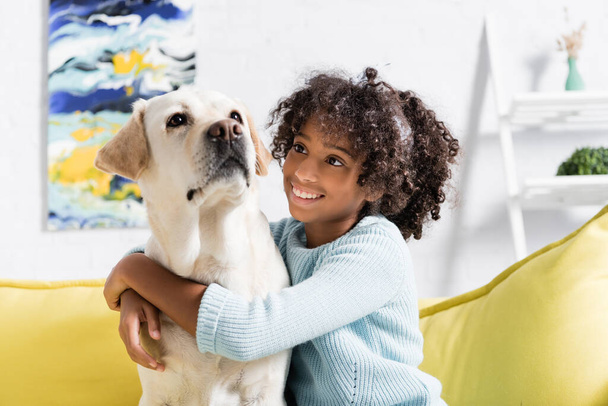 Chica afroamericana sonriente abrazándose y mirando al labrador, mientras está sentada en un sofá amarillo en casa, sobre un fondo borroso - Foto, Imagen