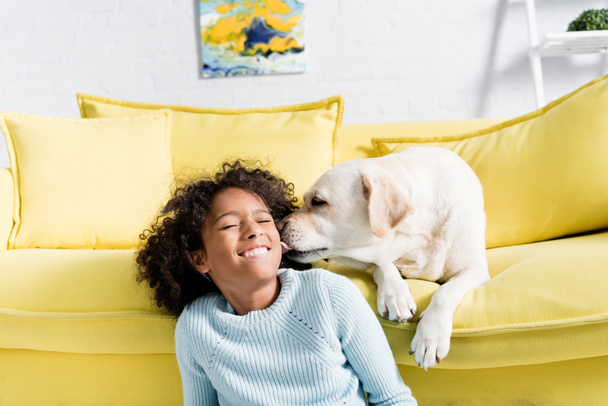 Labrador leckt Mädchen die Wange, während er zu Hause auf gelbem Sofa liegt - Foto, Bild