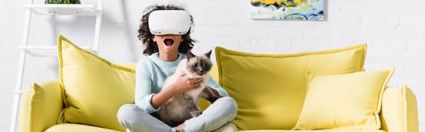 African american girl with open mouth in vr headset embracing cat, while sitting on sofa at home, banner - Photo, Image