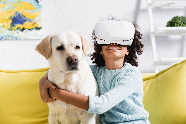 Smiling curly african american girl hugging retriever, while wearing vr headset, sitting on sofa on blurred background - Fotó, kép