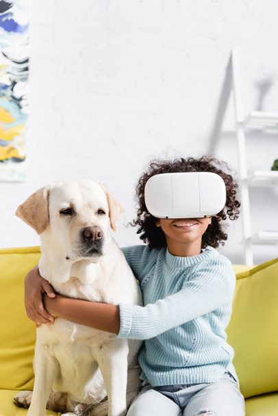 Happy curly african american girl hugging retriever, while wearing vr headset, sitting on sofa on blurred background - Fotografie, Obrázek
