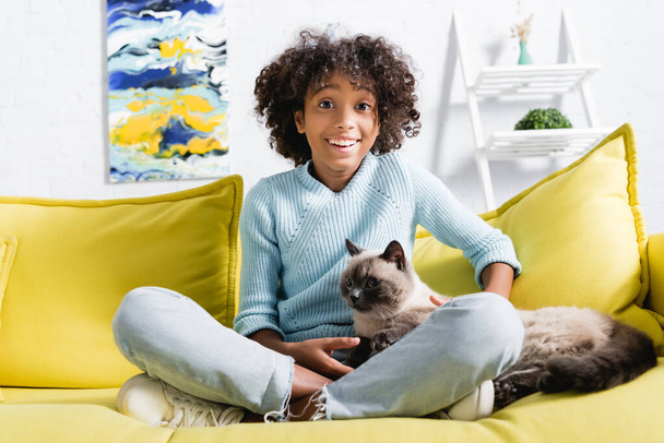 Happy african american girl with crossed legs looking at camera, while stroking cat on sofa on blurred background - Photo, Image