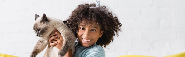 Cheerful african american girl holding siamese cat, while looking at camera at home, banner - Foto, Imagen