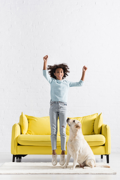 Happy african american girl with hands in air jumping on white rug near labrador at home - Photo, Image