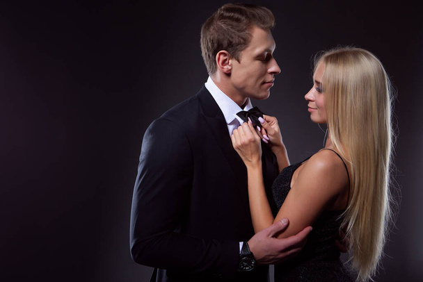 A charming blonde woman in a black evening dress lovingly ties a tie for her man. A young couple in love. - 写真・画像