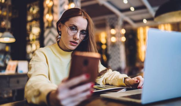 Caucasian hipster girl in spectacles reading received notification for cellphone application, millennial woman in classic eyewear using modern device during time for studying in coworking space - Fotografie, Obrázek