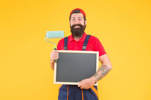 He loves his job. reconstruction advertising. creativity. happy man with paint roller. painter at painting works. hipster with copy space on blackboard. repair and building. craftsman on construction - Photo, Image