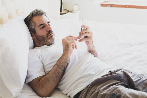 Der Mann liegt auf dem Bett und benutzt ein Handy. Ein Mann ruht sich aus und schreibt eine Nachricht auf sein Handy. - Foto, Bild